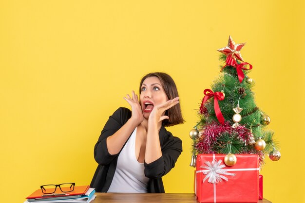 Shocked young woman looking at something sitting at a table near decorated Christmas tree at office on yellow 