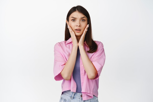 Shocked young woman looking worried holding hands near face with concerned emotion standing over white background