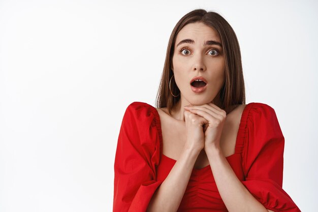 Shocked young woman looks startled and amazed, speechless staring at camera, open mouth and gasp worried, hear big news, standing against white background