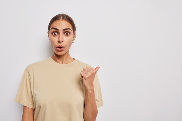 Shocked young woman points thumb away shows special sale or price discount keeps jaw dropped wears casual beige t shirt isolated over white wall impressed to tell about promotional offer