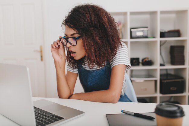 Shocked young woman in striped shirt looking at laptop screen and holding glasses