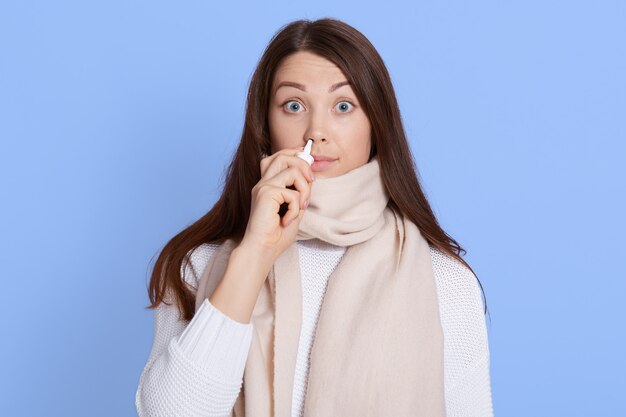 Shocked young woman in white sweater and scarf holding and using nasal drops with big eyes isolated on blue wall wall, dark haired lady feels bad, suffering from runny nose, catching cold.