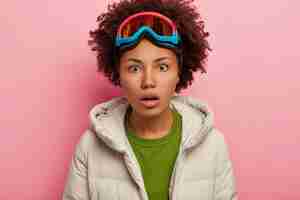 Free photo shocked young woman in winter clothes, wears snowboarding mask on head, holds breath from wonder, isolated over pink background