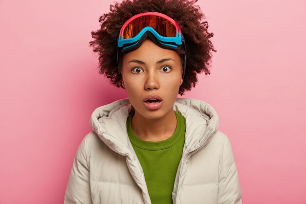 Шокированная молодая женщина в зимней одежде, носит маску для сноубординга на голове, задерживает дыхание от чуда, изолирована на розовом фоне