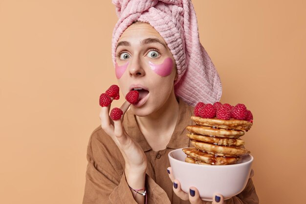 Shocked young woman with raspberries on fingers looks surprisingly at camera has delicious breakfast holds bowl of pancakes with syrup applies beauty pads under eyes wears wrapped towel on head