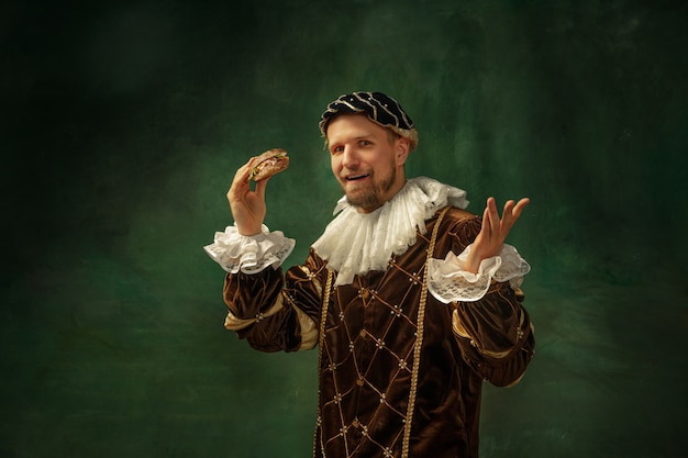 Shocking food. Portrait of medieval young man in vintage clothing with wooden frame on dark background. Male model as a duke, prince, royal person. Concept of comparison of eras, modern, fashion.