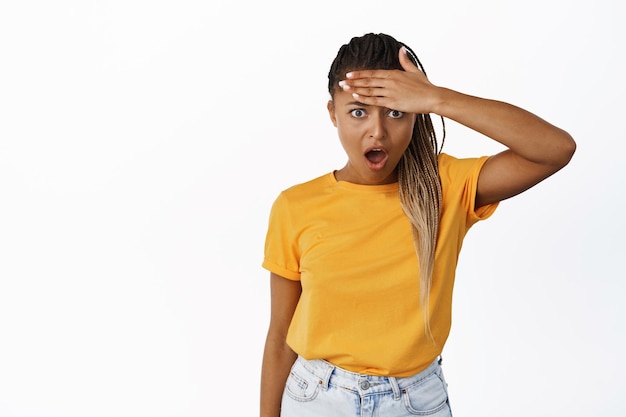 Shocking news Speechless girl slap forehead and drop jaw staring shocked at camera standing i yellow tshirt over white background