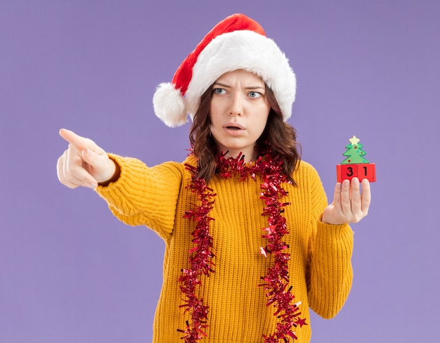 Shoked young slavic girl with santa hat and with garland around neck holds christmas tree ornament looking and pointing at side isolated on purple background with copy space