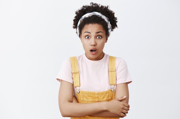 Shook impressed African American girl in headband and yellow overalls, holding hands together on chest and gazing