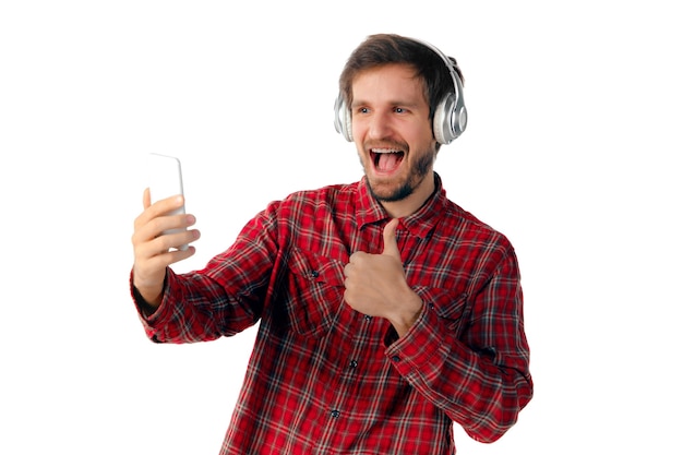 Shoot of young caucasian man using mobile smartphone, headphones isolated on white studio background. Concept of modern technologies, gadgets, tech, emotions