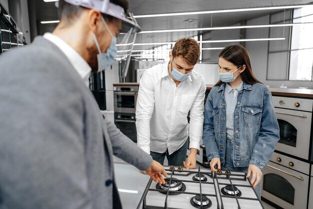 Shop assistant in hypermarket shows new model of gas stove to young couple all wearing medical masks