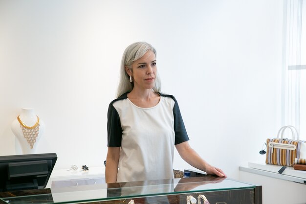 Shop cashier or seller working in trendy boutique, standing at desk with cash register and looking away. Medium shot. Retail store or job concept