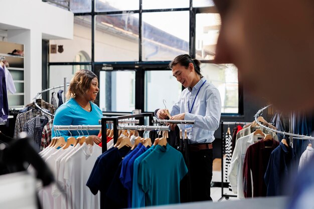 新しいファッション コレクションを見て、衣料品店で服の素材について話し合う買い物中毒の顧客。アフリカ系アメリカ人の女性がフォーマルウェアを買い、スタイリッシュなアイテムが入ったラックをチェックする