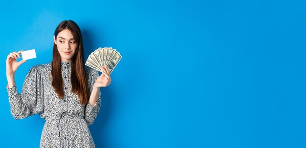 Shopping beauty girl in dress looking aside and smiling pensive showing money and plastic credit car