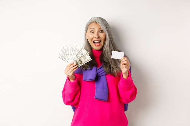 Shopping concept. Amazed asian granny showing plastic credit card and money dollars, want to buy something, standing over white background.