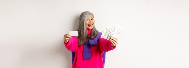 Free photo shopping concept cheerful asian senior woman showing cash and plastic credit card smiling at camera