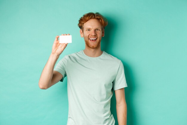 ショッピングのコンセプト。プラスチック製のクレジットカードを示し、ミントの背景の上に立って微笑んで、Tシャツを着た陽気な若い男。