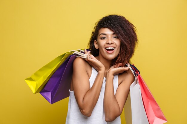 쇼핑 개념-가까이 초상화 젊은 아름 다운 매력적인 아프리카 여자 웃 고 화려한 쇼핑 가방과 함께 즐거운. 노란 파스텔 벽 배경입니다. 공간을 복사하십시오.