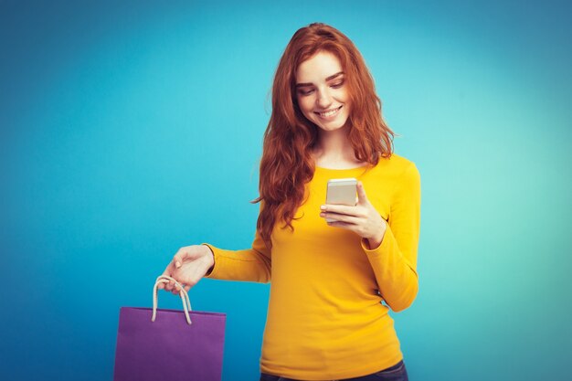 ショッピングコンセプト - クローズアップ肖像画若い美しい魅力的な赤毛の女の子ショッピングバッグでカメラを見て笑って。青いパステルの背景。スペースをコピーします。