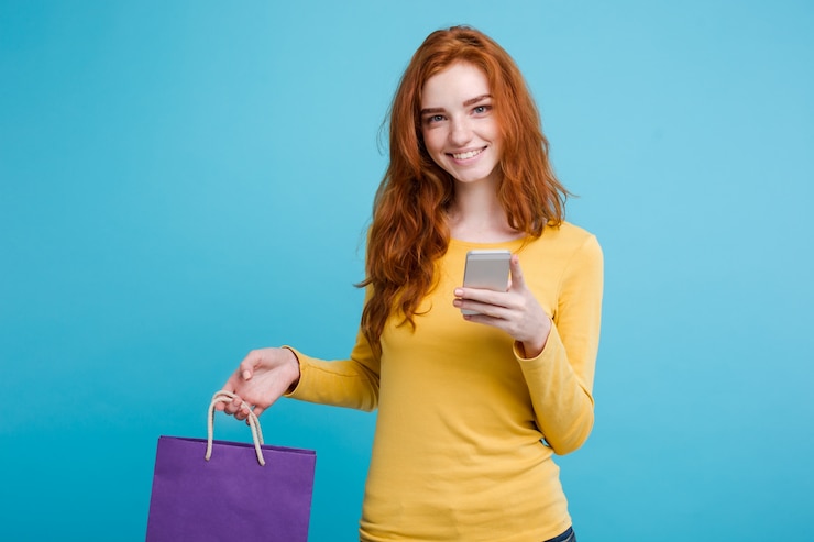 Free Photo shopping concept - close up portrait young beautiful attractive redhair girl smiling looking at camera with shopping bag. blue pastel background. copy space.