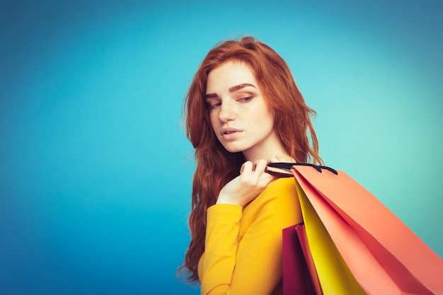 ショッピングコンセプト - クローズアップ肖像画若い美しい魅力的な赤毛の女の子ショッピングバッグでカメラを見て笑って。青いパステルの背景。スペースをコピーします。
