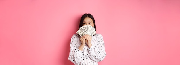 Free photo shopping concept cute asian woman hiding face behind money dollars peeking at camera standing over p