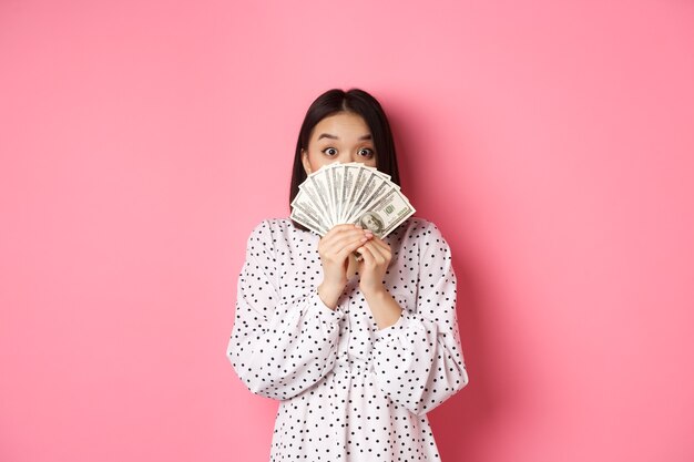 Shopping concept cute asian woman hiding face behind money dollars peeking at camera standing over p...