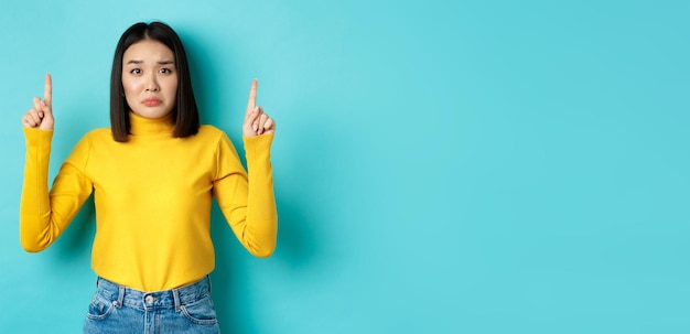 Free photo shopping concept disappointed and gloomy asian woman sulking upset pointing fingers up at bad news b