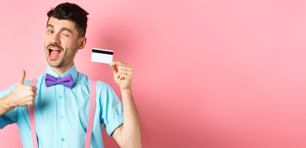 Foto gratuita concetto di shopping ragazzo festivo in cravatta a farfalla che fa l'occhiolino alla macchina fotografica che mostra i pollici in su con la carta di credito in plastica