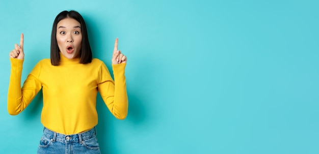 Free photo shopping concept impressed young korean woman showing advertisement on top pointing fingers up and g