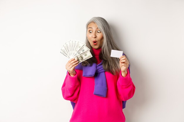 Shopping concept. Lucky asian senior woman looking amazed at money and showing plastic credit card, standing over white background