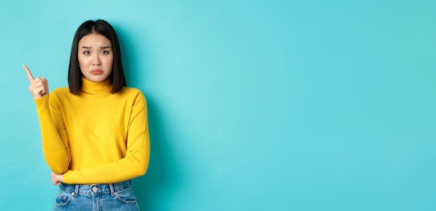 Free photo shopping concept sad and gloomy asian girl in yellow sweater pointing finger left frowning and showi