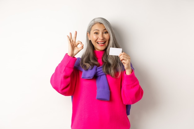 Shopping concept. Smiling asian middle-aged woman with grey hair showing plastic credit card and OK sign, recommending bank promotion, white background