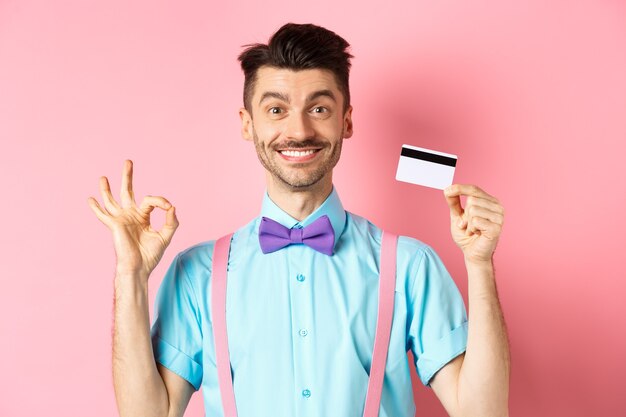 Shopping concept. Smiling handsome male shopper showing Ok sign and plastic credit card, buying something, standing satisfied on pink background.