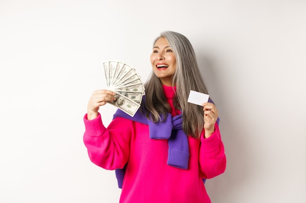 Free photo shopping concept. stylish asian senior woman showing money dollars and plastic credit card, looking dreamy aside, thinking about buying, white background.