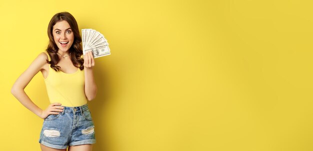 クレジットとお金の概念をショッピング若いブルネットの女性現金を示し、黄色の背景の上に立って喜んで笑顔