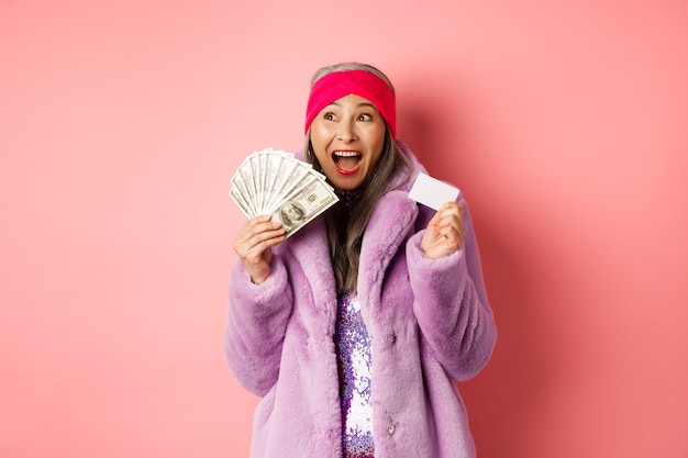 ショッピングとファッションのコンセプト。アジアの年配の女性は勝者のように幸せに叫び、ドルのお金とプラスチックのクレジットカードを持って、興奮しているように見える、ピンクの背景