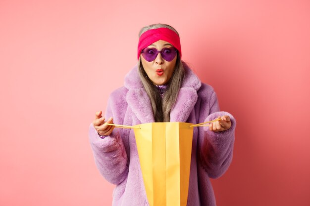 Шоппинг и концепция моды. Стильная азиатская пожилая женщина в солнцезащитных очках и шубе из искусственного меха открывает бумажный пакет с подарками, удивленно глядя в камеру, розовый фон