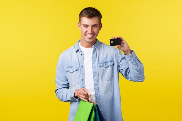 Shopping, leisure and discounts concept. Smiling handsome young man buying new clothes, holding bags and showing credit card with satisfied expression, paying with saved money, yellow background.