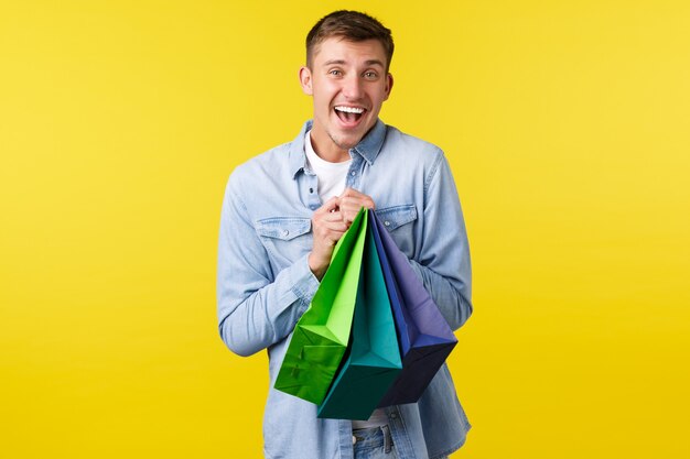 ショッピング、レジャー、割引のコンセプト。幸せなハンサムな男の買い物好きな笑顔、特別オファーで物を買うのが好き、満足のいく表情、黄色の背景のバッグを持っています。