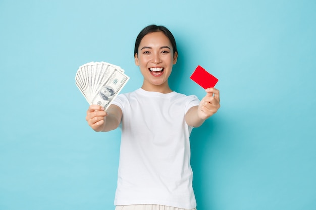 쇼핑, 돈 및 금융 개념. 밝은 파란색 벽 위에 만족 서 자랑스러운 표정으로 현금 및 신용 카드에 달러를 보여주는 행복 하 고 기쁘게 웃는 아시아 소녀.