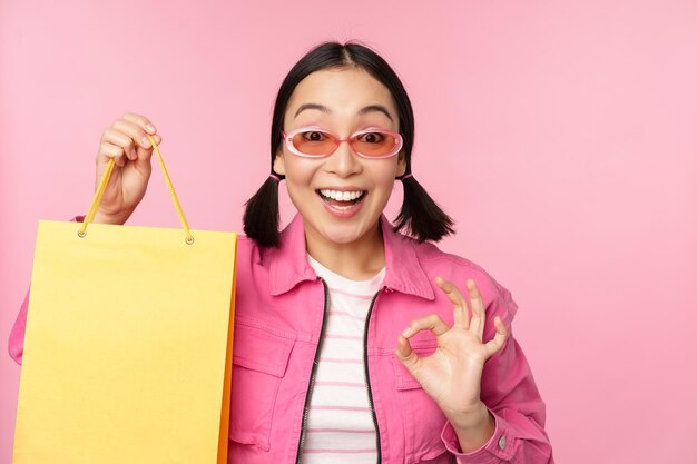 선글라스를 끼고 쇼핑하는 세련된 아시아 소녀는 상점에서 가방을 보여주고 분홍색 배경 위에 서 있는 상점에서 판매 프로모션을 추천하며 웃고 있습니다.