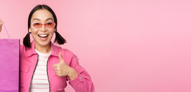 Shopping elegante ragazza asiatica in occhiali da sole che mostra la borsa dal negozio e sorridente consigliando promo di vendita in negozio in piedi su sfondo rosa