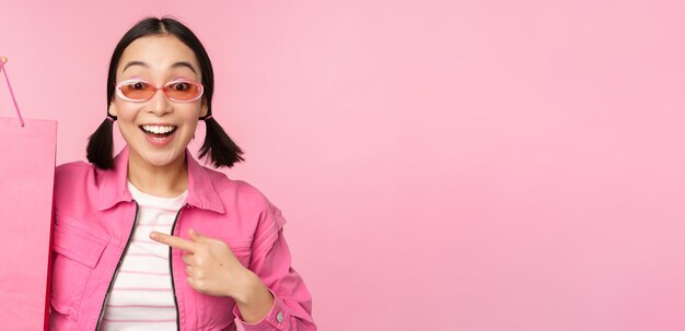 Shopping Stylish asian girl in sunglasses showing bag from shop and smiling recommending sale promo in store standing over pink background
