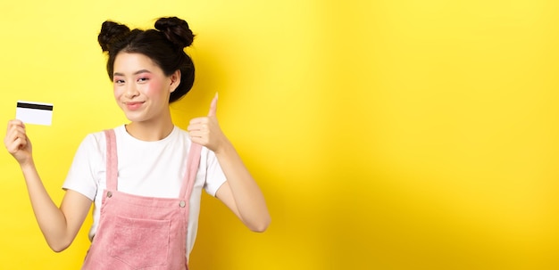 Free photo shopping stylish asian glam girl showing thumb up and plastic credit card standing on yellow backgro