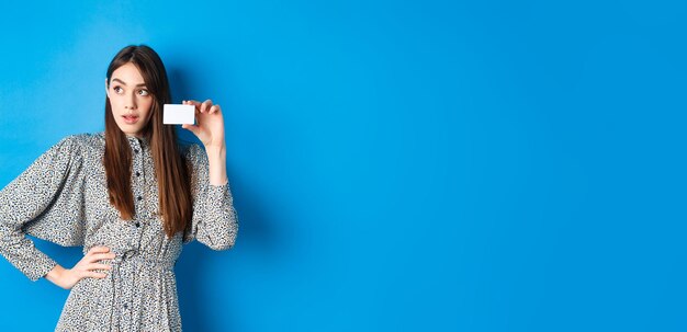 Shopping thoughtful female shopper looking left at logo showing plastic credit card standing on blue