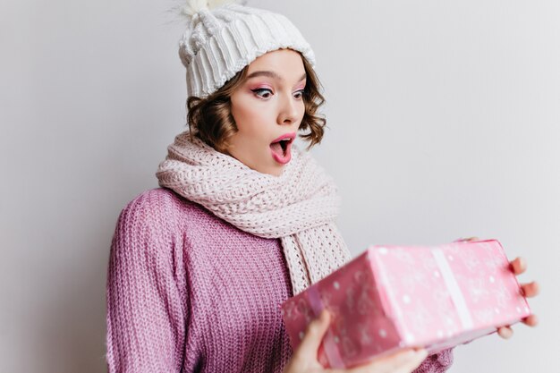 Short-haired european girl in hat posing emotionally, looking at new year present. Indoor photo of amazed young lady in cute scarf holding pink gift.