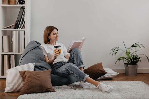 Short-haired girl in white T-shirt with smile reading book while sitting on soft bean bag.