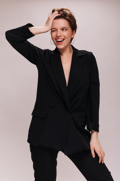 Short haired lady in black outfit winking on white background. Charming caucasian woman in dark suit widely smiling on isolated