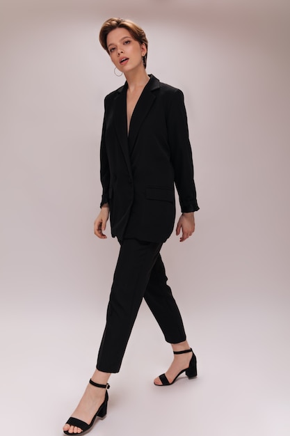 Short haired lady in dark suit moving on white background. Charming woman in black pamts and oversize jacket walks on isolated